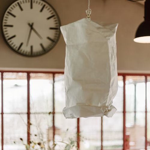 A white paper ceiling lamp is shown hanging as a pendant in a home setting, with a window and a clock in the background.