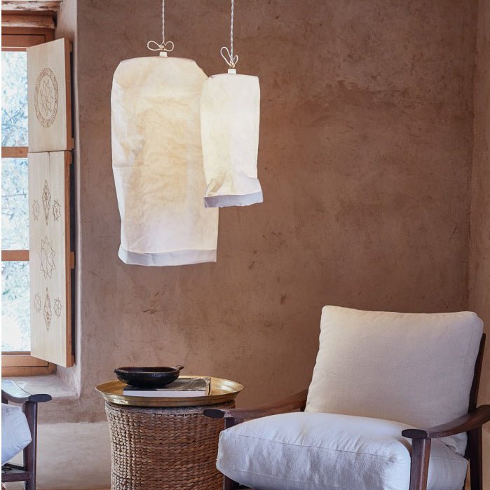 Two white washable paper lamps are shown hanging from a ceiling in varying sizes. The background shows a raw plaster wall, a white armchair and a raffia side table holding a gold tray.