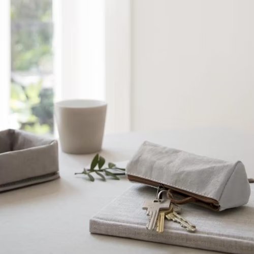 A grey washable paper key holder is shown atop a book. At left sits a grey washable paper tray, and in the background sits a white ceramic cup.