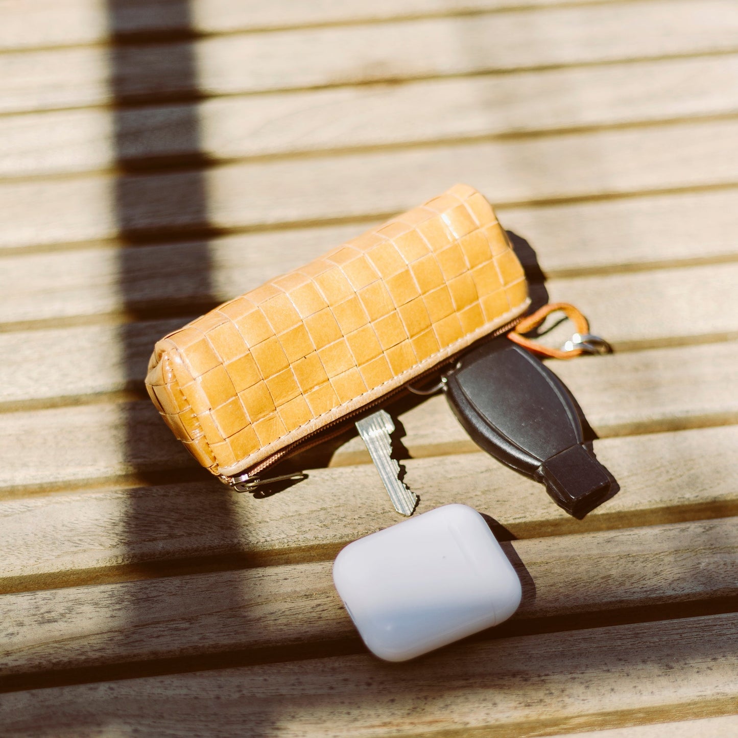 A tan woven washable paper key holder is shown on wooden slats. It is unzipped to reveal a house key, a car key, and a set of Air Pods.