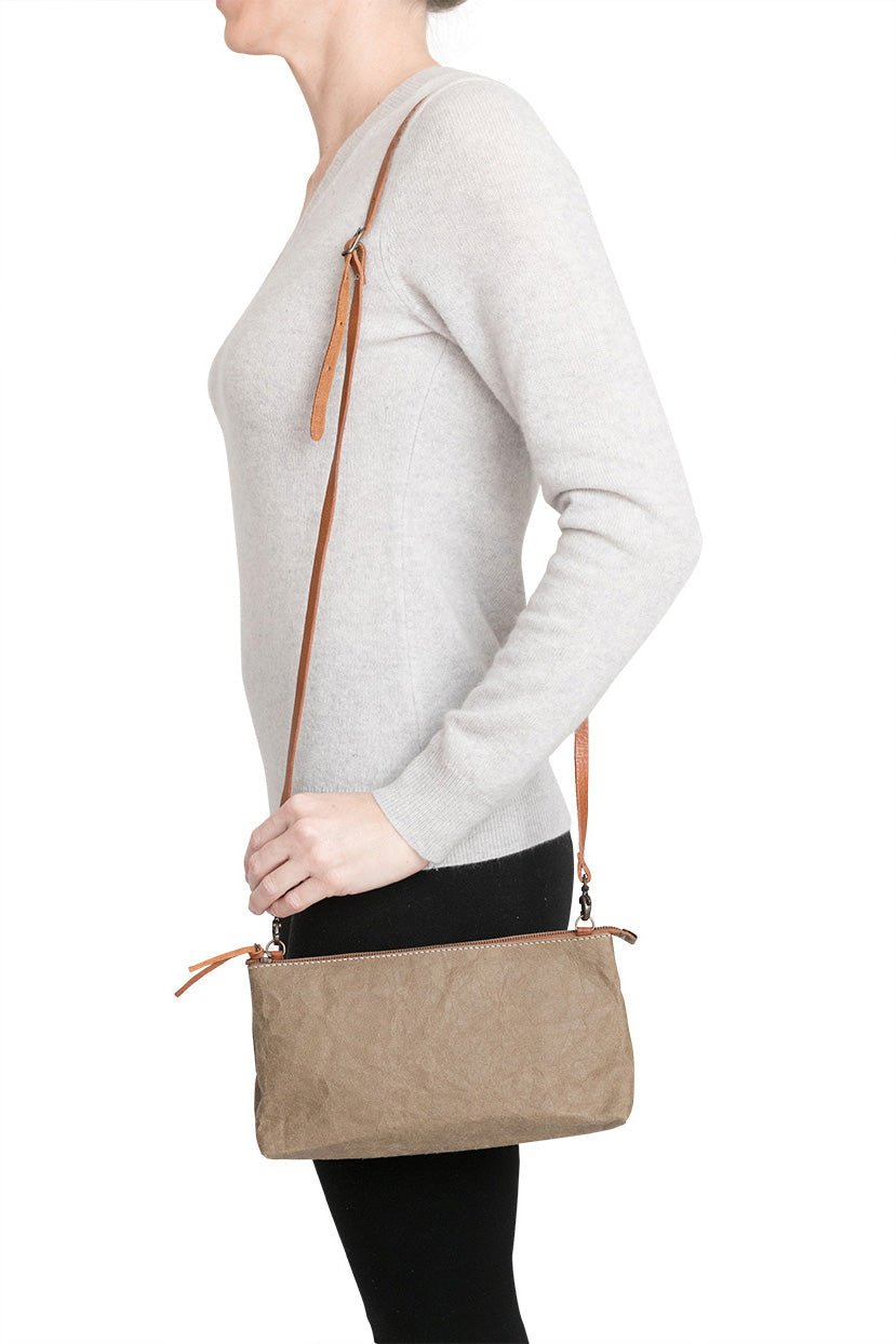 A woman is shown smiling in casual clothing, wearing a small washable paper handbag in pale brown, with a tan long strap.