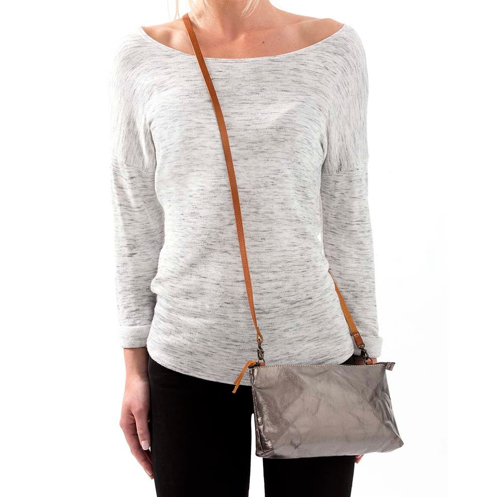 A woman is shown in casual clothing wearing a metallic pewter washable paper small handbag in a cross-body style, by way of a long tan strap.