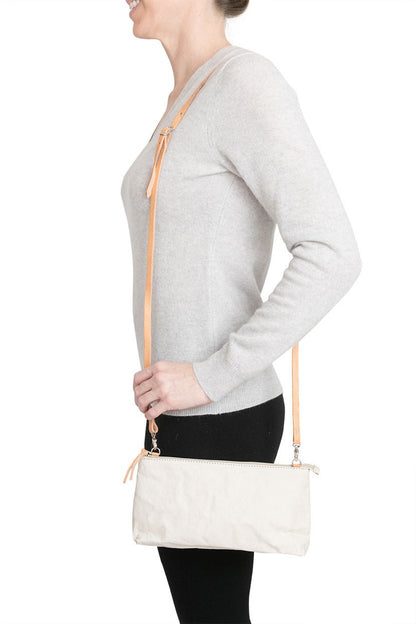 A woman is shown smiling in casual clothing, wearing a small washable paper handbag in white, with a tan long strap.
