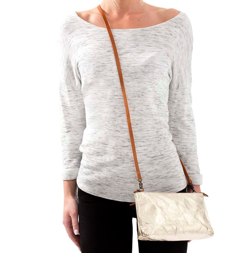 A woman is shown in casual clothing wearing a metallic gold washable paper small handbag in a cross-body style, by way of a long tan strap.