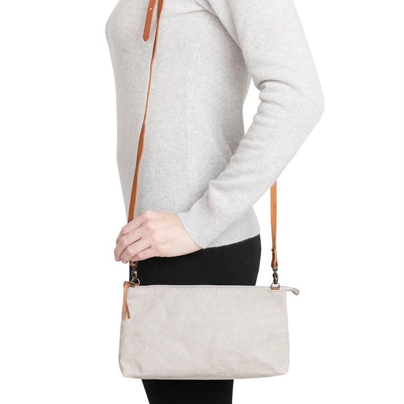 A woman is shown smiling in casual clothing, wearing a small washable paper handbag in grey, with a tan long strap.