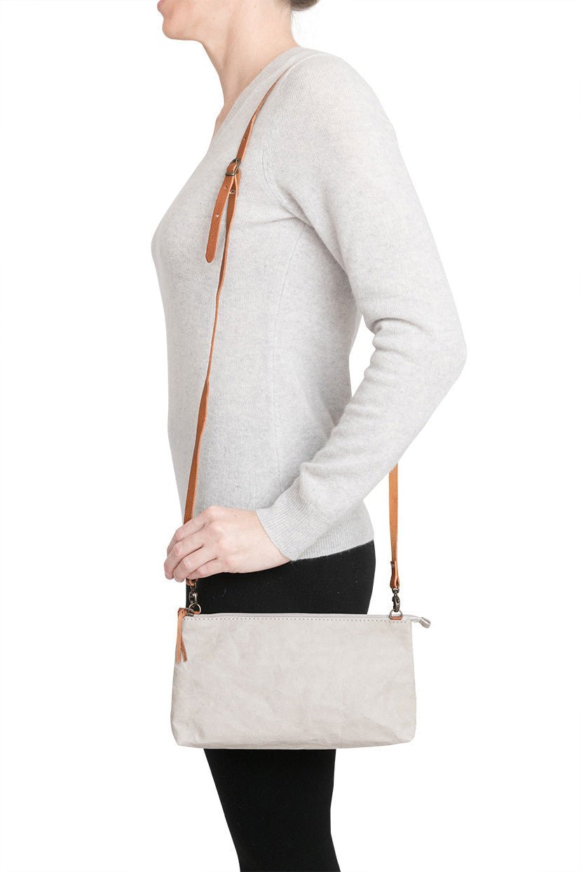 A woman is shown smiling in casual clothing, wearing a small washable paper handbag in grey, with a tan long strap.