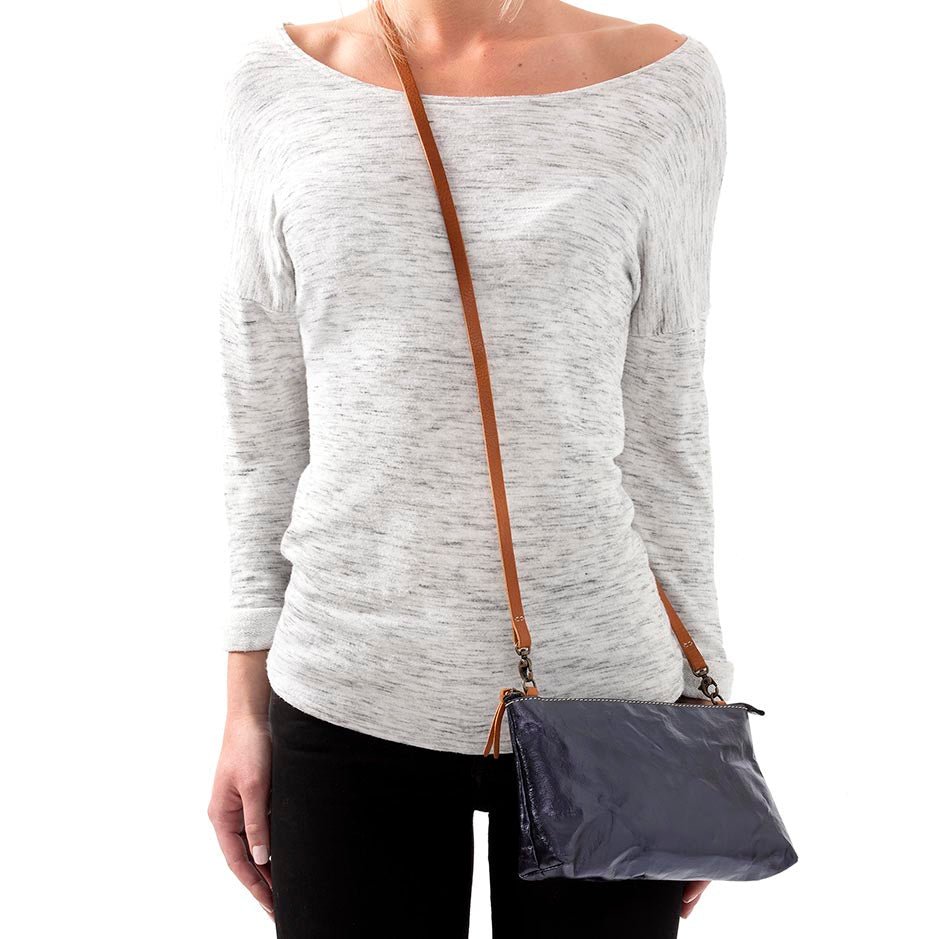 A woman is shown in casual clothing wearing a metallic navy washable paper small handbag in a cross-body style, by way of a long tan strap.