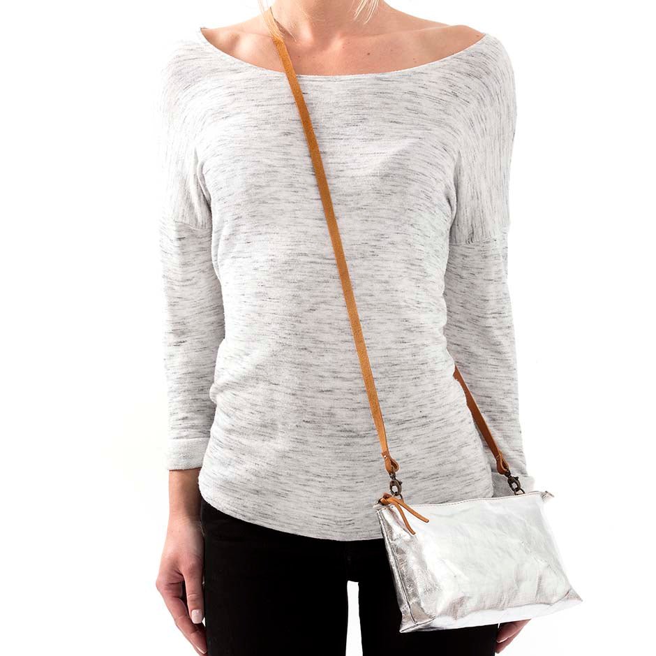 A woman is shown in casual clothing wearing a metallic silver washable paper small handbag in a cross-body style, by way of a long tan strap.