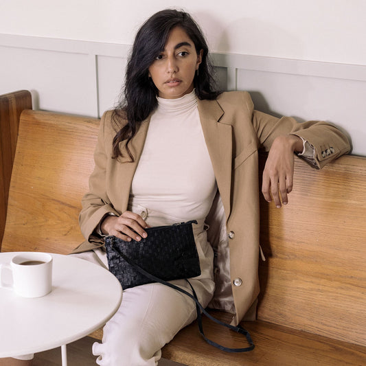 A woman is shown at a restaurant table wearing a camel jacket and white trousers. On her lap sits a black woven washable paper small handbag with a long black strap.