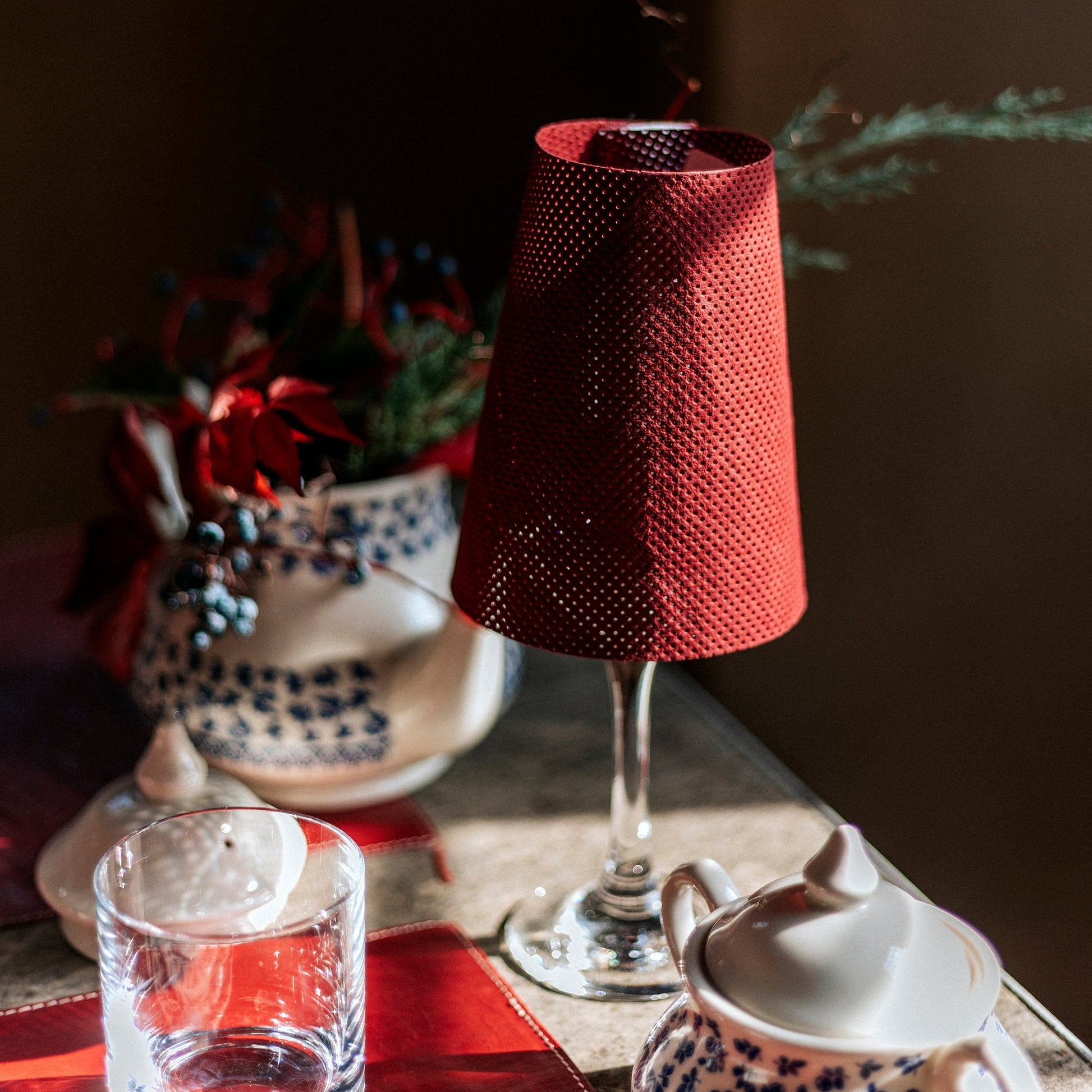 A red perforated washable paper lampshade is shown sitting atop a wine glass in a Christmas dinner party setting.