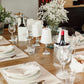 A series of white washable paper lampshades with cut out details sit atop wine glasses in a dinner party setting. A silver metallic and a white washable paper wine cooler hold wine bottles, and washable paper placemats feature on the table.