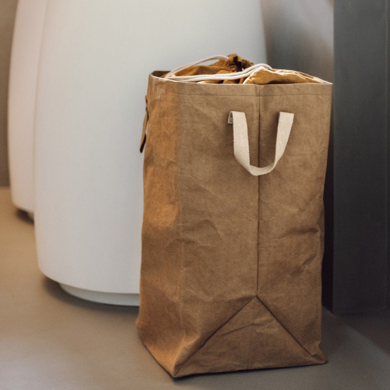 A washable paper laundry bag is shown in tan in a home setting, from a side angle.