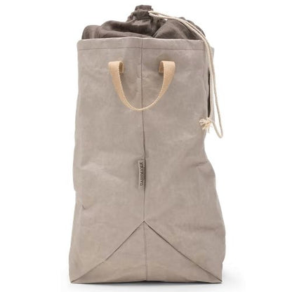 A washable paper laundry bag is shown in beige from a side angle. It features two side handles, a popper tab, and an interior drawstring lining.