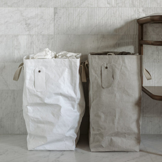 Two washable paper laundry bags sit side by side in a marble home setting. The one at left is white and the one at right is grey.