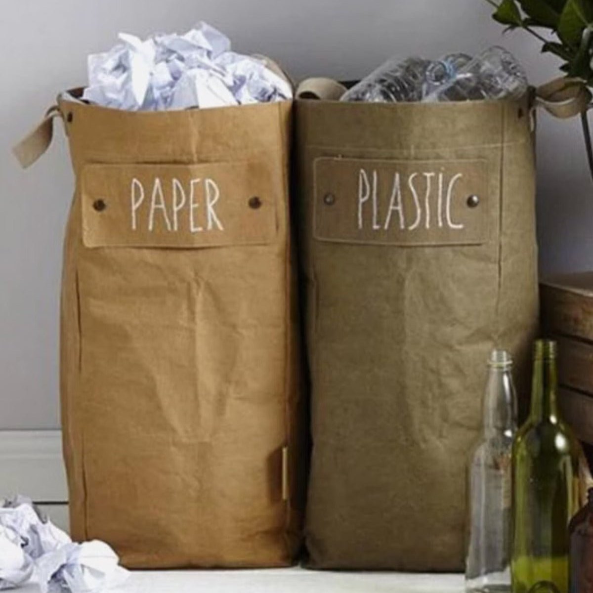 Two washable paper tall bags sit nestled together by popper closures, in a home setting. The one at left is tan and labelled "paper" and the one at right is khaki and labelled "plastic."