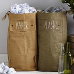 Two washable paper tall bags sit nestled together by popper closures, in a home setting. The one at left is tan and labelled "paper" and the one at right is khaki and labelled "plastic."