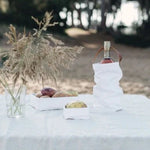 An outdoor table setting shows a vase of flowers, a large washable paper tray containing potatoes, a small washable paper tray containing a vegetable, and a white washable paper wine bag sits at right, holding a bottle of wine. 