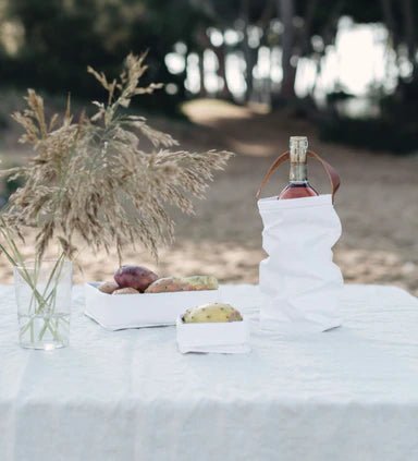 An outdoor table setting shows a vase of flowers, a large washable paper tray containing potatoes, a small washable paper tray containing a vegetable, and a white washable paper wine bag sits at right, holding a bottle of wine. 