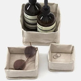 A set of three washable paper trays are shown in grey, in varying sizes. The largest contains two bottles of handsoap, the medium contains a pair of sunglasses, and the smallest contains a pair of earrings.