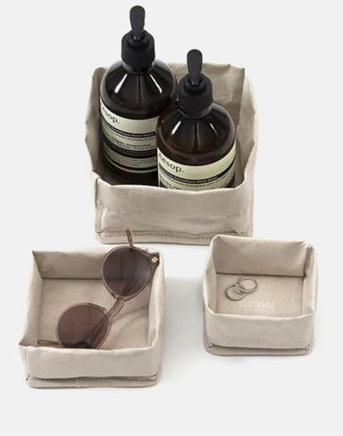 A set of three washable paper trays are shown in grey, in varying sizes. The largest contains two bottles of handsoap, the medium contains a pair of sunglasses, and the smallest contains a pair of earrings.