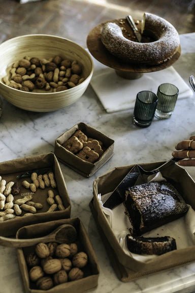 A series of washable paper trays adorn a marble table top. They contain walnuts, peanuts, biscuits, and a cake. They are all of varying sizes.