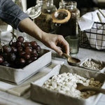 A person's hand reaches across a marble table top, displaying washable paper trays in grey of varying sizes. They contain grapes and nuts, and have been used as serve wear. 