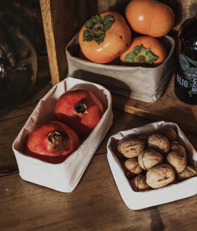 A set of three washable paper trays in grey sit on a wooden table. All in varying sizes, they contain pomegranates and walnuts.
