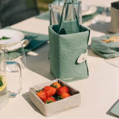 A washable paper tray in grey containing strawberries sits atop an outdoor table setting. Behind it sits a sage green washable paper cooler containing a glass water bottle.