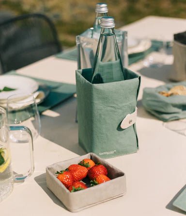 A washable paper tray in grey containing strawberries sits atop an outdoor table setting. Behind it sits a sage green washable paper cooler containing a glass water bottle.
