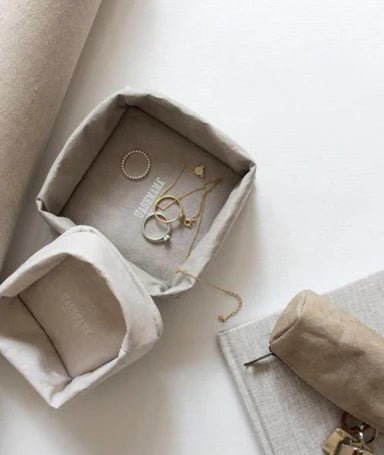 Two washable paper trays lie one on top of the other. The top one is small and cream, the bottom one is larger and contains jewellery. A washable paper key holder sits at right on top of a book. 