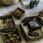 A series of washable paper trays adorn a marble table top. They contain walnuts, peanuts, biscuits, and a cake. They are all of varying sizes.