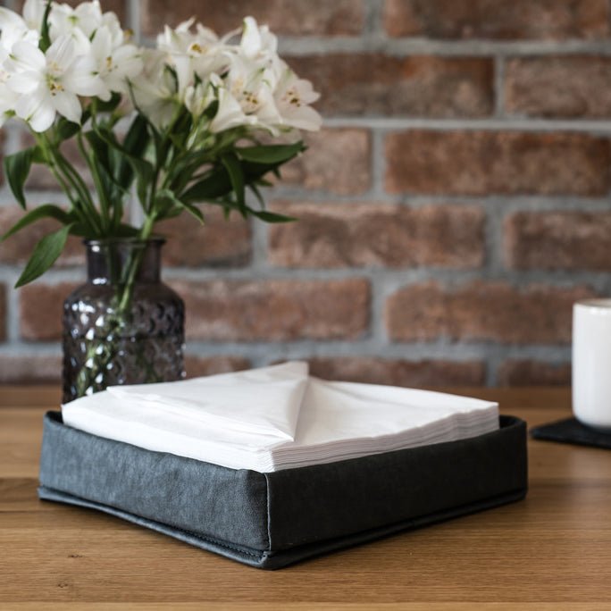 A black washable paper tray is shown containing napkins on a tabletop setting, with a vase containing white lilies in the background.