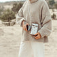 A woman is shown on the beach wearing a beanie hat, a beige jumper and white trousers. She is wearing a washable paper phone pouch crossbody in metallic silver.