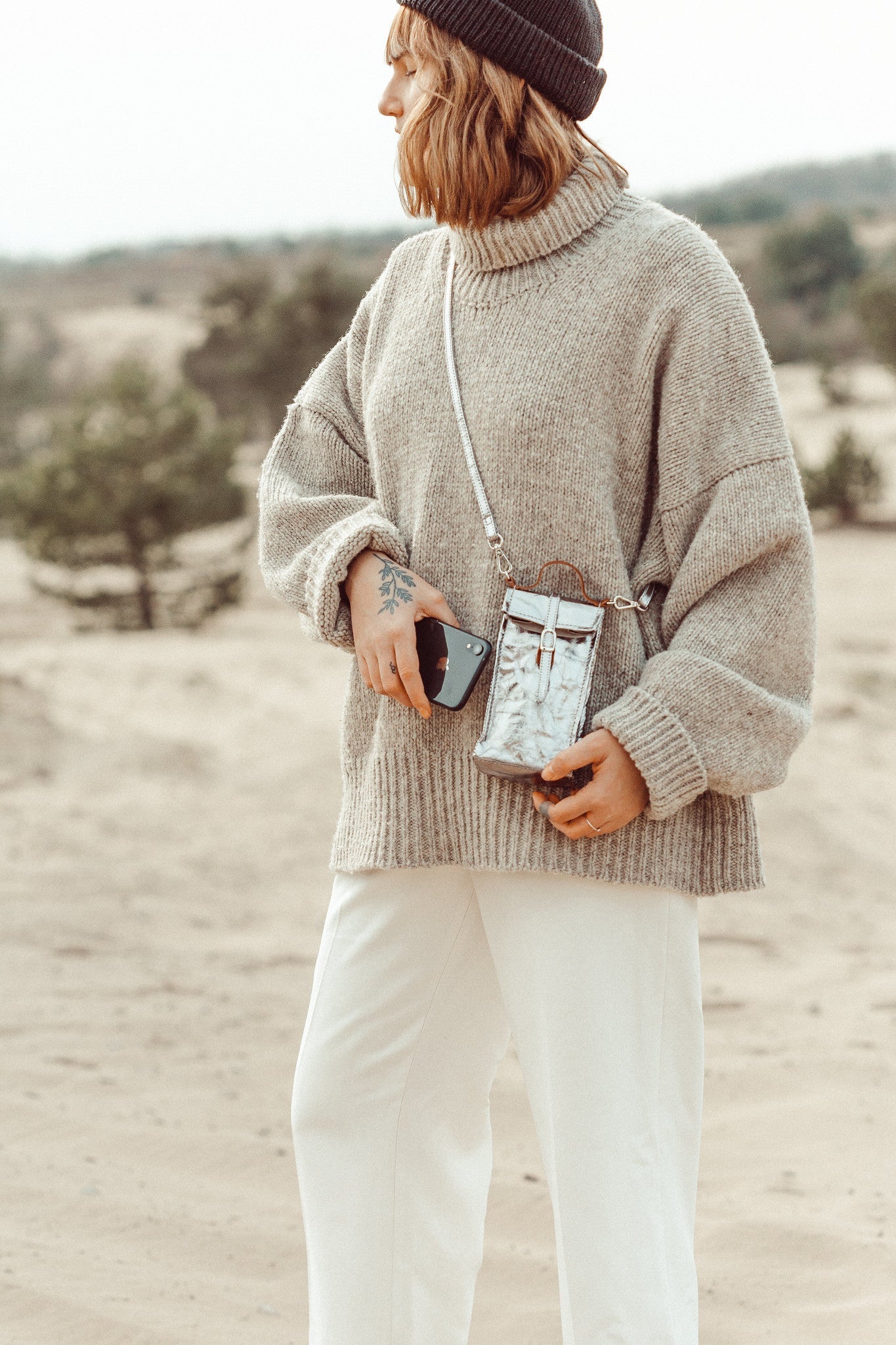 A woman is shown on the beach wearing a beanie hat, a beige jumper and white trousers. She is wearing a washable paper phone pouch crossbody in metallic silver.