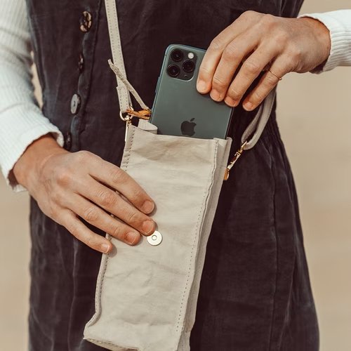 A woman is shown wearing a grey washable paper phone pouch crossbody, on a long washable paper strap. She is pulling an iPhone out of it with her left hand and holding the pouch with her right hand.