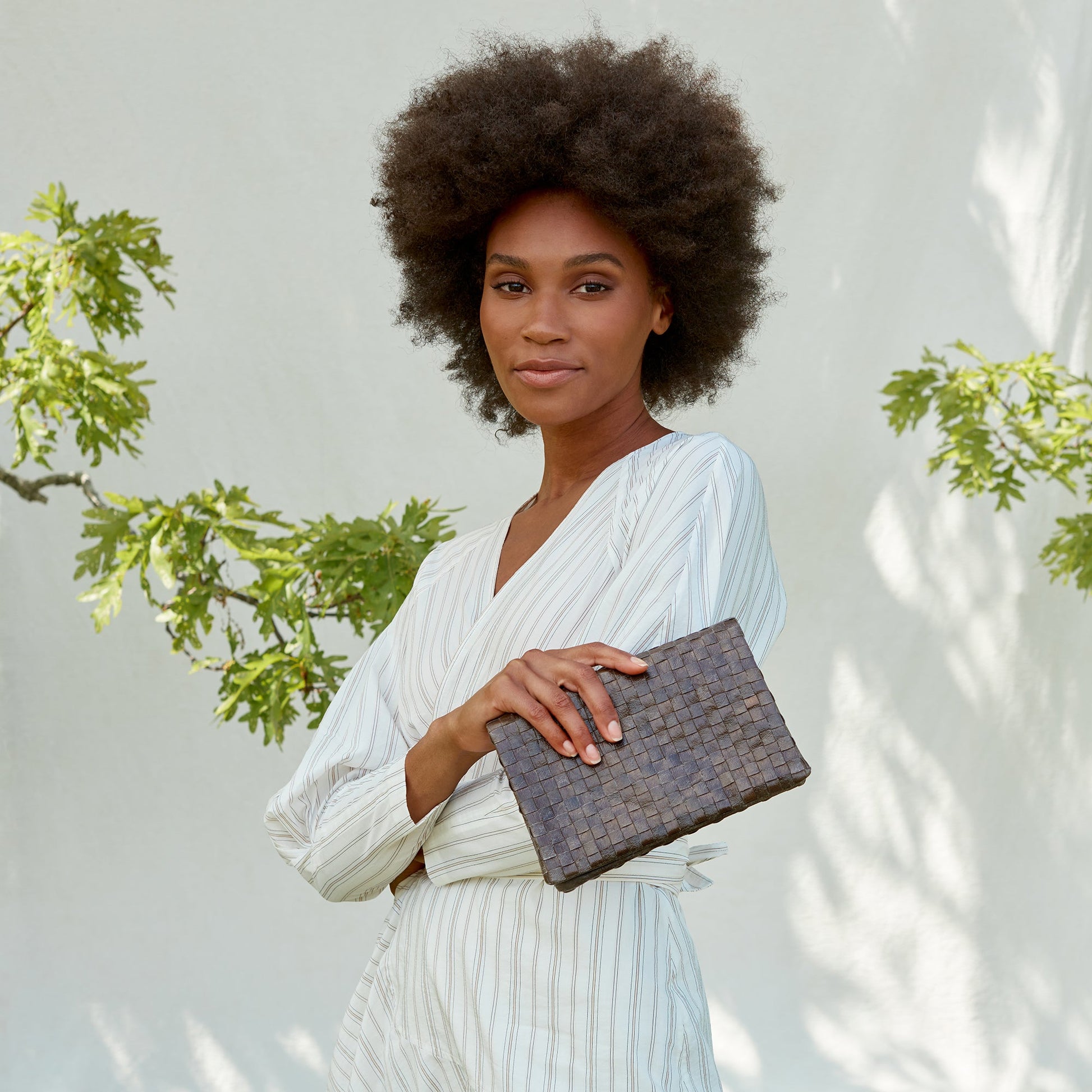 A woman is shown standing outside, amongst foliage, wearing white. She is holding a chocolate brown woven washable paper clutch bag. 