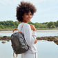 A woman is shown by a lake, wearing casual clothing. She is carrying a pewter metallic rounded washable paper backpack over one shoulder, by way of a canvas cream strap.
