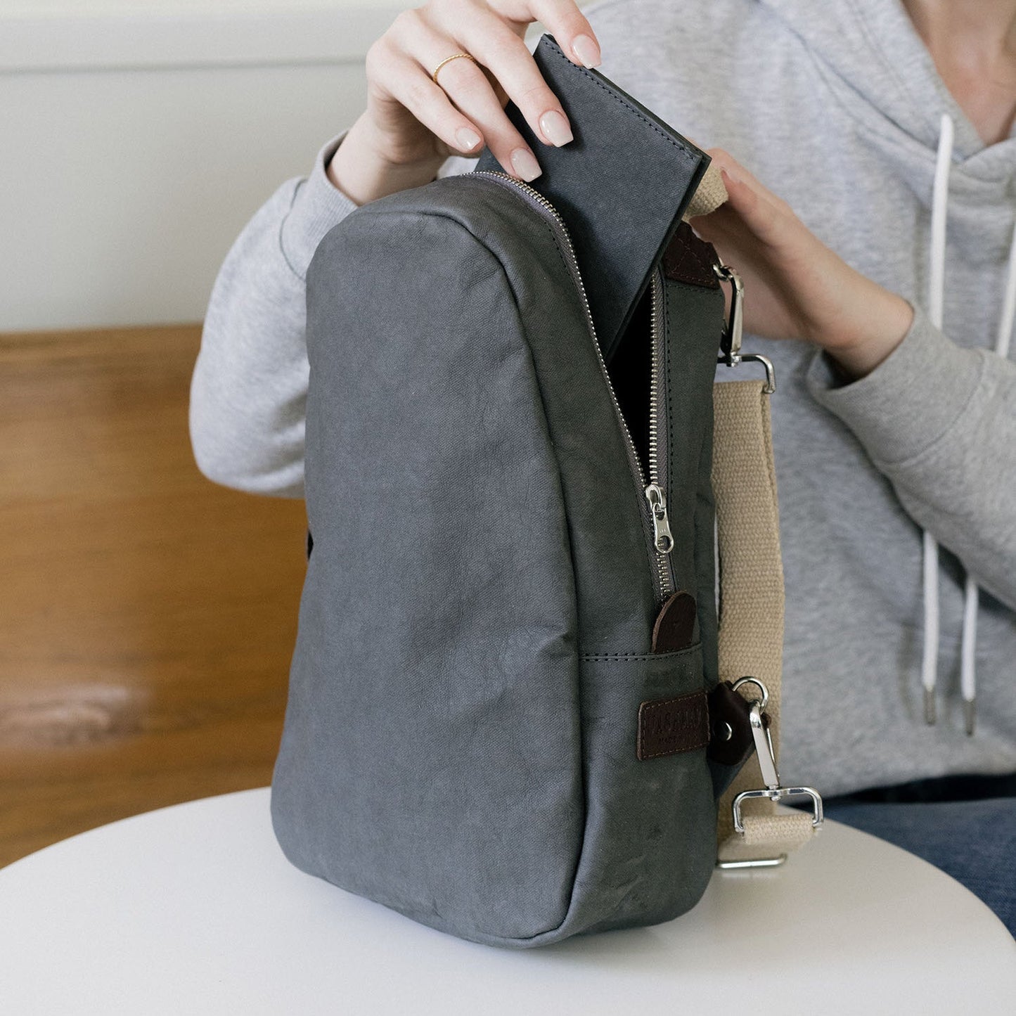 A woman is shown wearing a grey hoodie, seated at a table. On the table sits a grey washable paper backpack with a silver zip, a brown side tab, and natural toned cotton canvas straps. The woman is placing a washable paper pouch into the backpack.