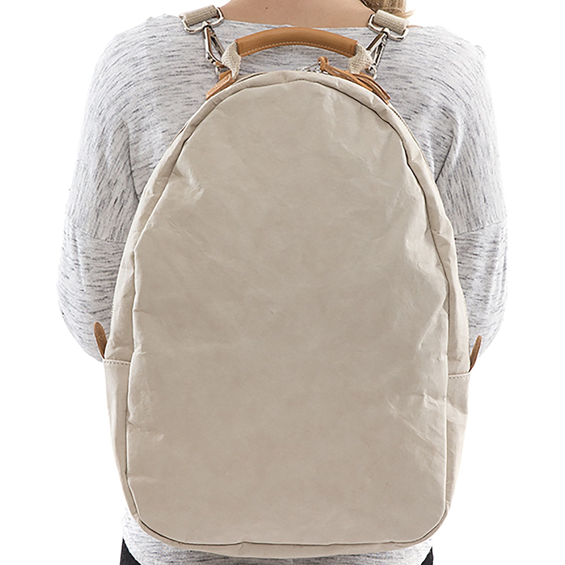 A blonde woman is shown wearing a washable paper backpack. It has two side zip toggles, is oval in shape and it is cream in colour.
