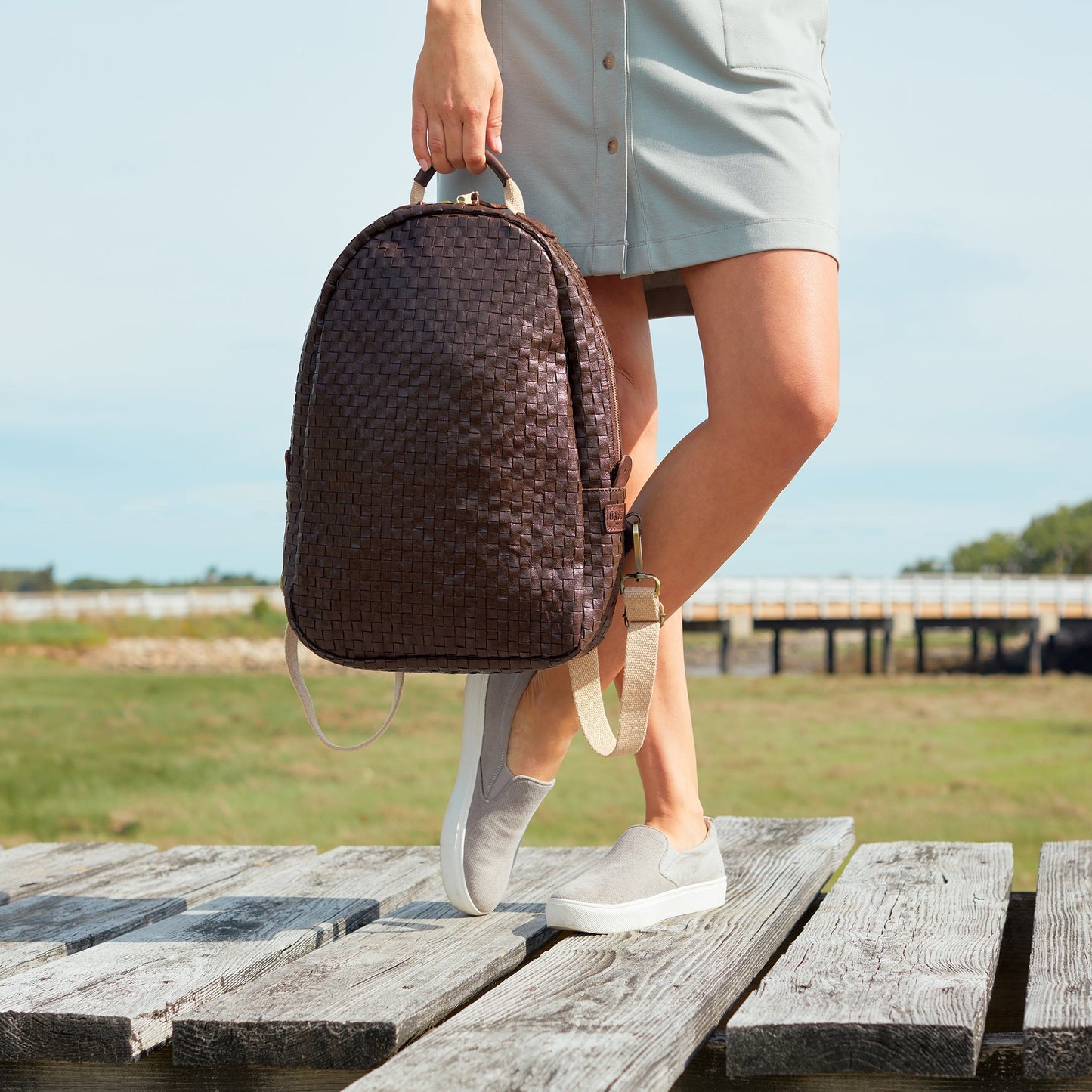 A woman is shown standing on decking, wearing a dress and plimsolls. In her right hand she holds a chocolate brown washable paper backpack by its top handle. 