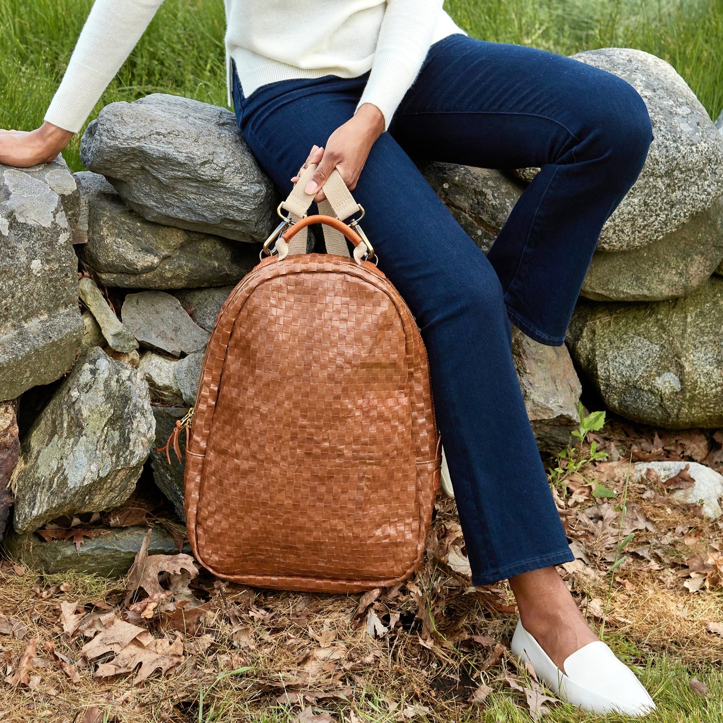 A woman is shown sitting on a stone wall, wearing jeans and a cream jumper. In her right hand she is holding a tan coloured washable paper woven backpack by its shoulder straps.