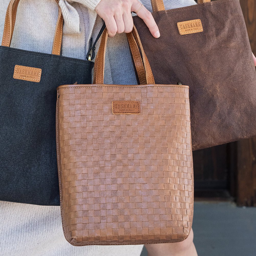 A woman is shown carrying three washable paper tote bags. From left to right they are black, woven tan, and chocolate brown, all shown from a close up angle.