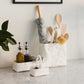 Two white washable paper utensil holders sit atop a marble counter. The larger contains a rolling pin and wooden spoons, the smaller contains salt and pepper. A white washable paper tray holds oil and vinegar at the side.