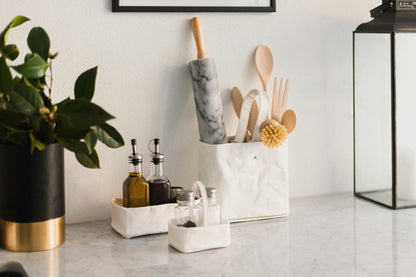 Two white washable paper utensil holders sit atop a marble counter. The larger contains a rolling pin and wooden spoons, the smaller contains salt and pepper. A white washable paper tray holds oil and vinegar at the side.