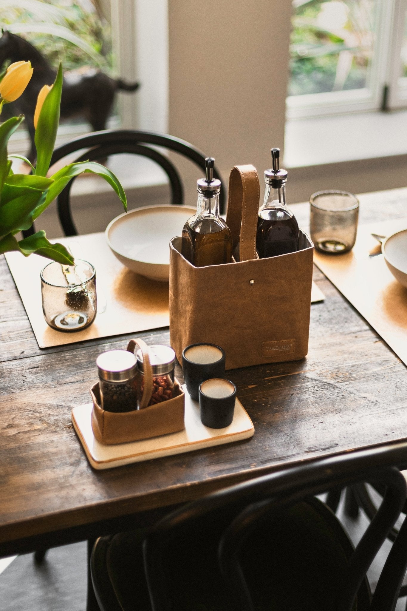 A wooden table top is set for a meal. A tan washable paper utensil holder contains an oil and vinegar bottle. A smaller washable paper utensil holder contains spices.