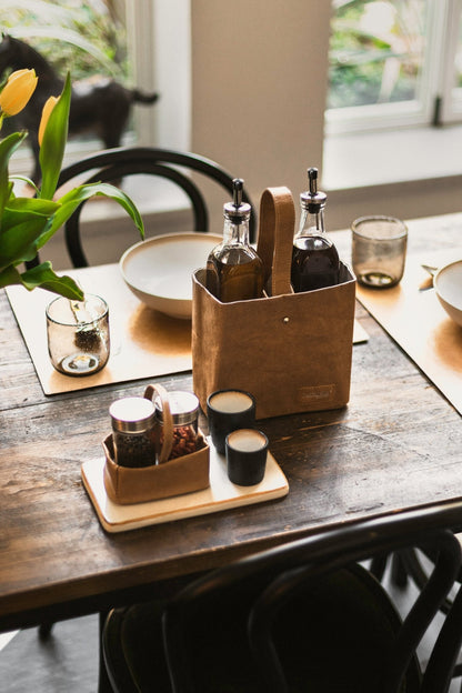 A wooden table top is set for a meal. A tan washable paper utensil holder contains an oil and vinegar bottle. A smaller washable paper utensil holder contains spices.