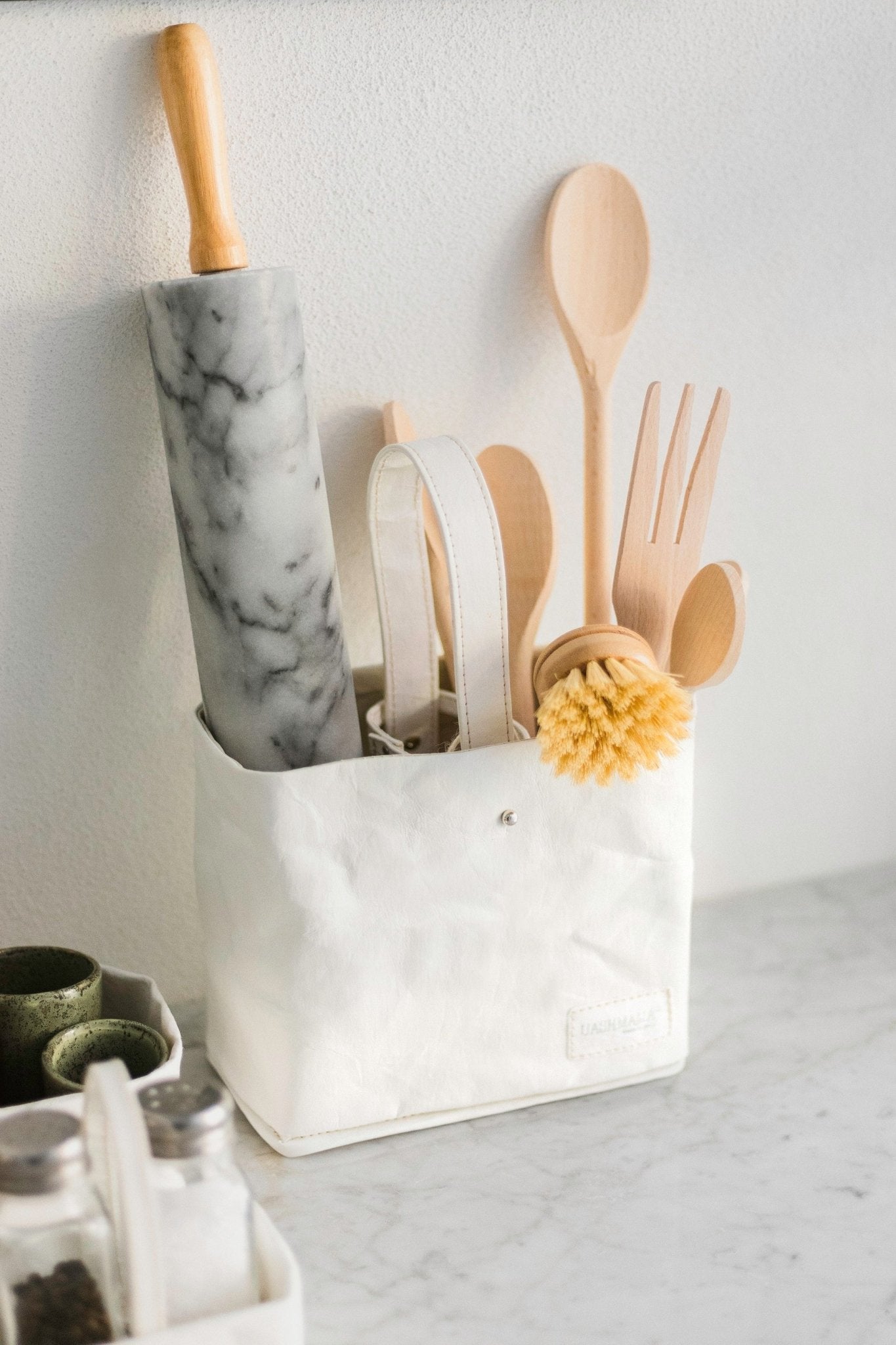A white washable paper utensil holder sits on a marble counter. It contains a marble rolling pin, wooden spoons and a dish brush.