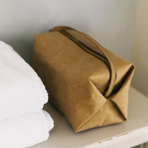 A tan washable paper origami pouch is shown on a shelf from a side angle, with a top handle. There is a stack of towels at left.