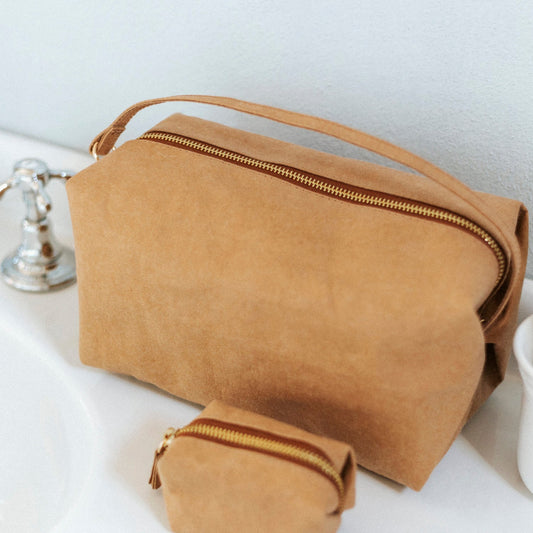 A large washable paper zip-top rectangular toiletry bag is shown atop a sink. It features a top handle, and there is a smaller washable paper pouch in front of it. They are both tan in colour.