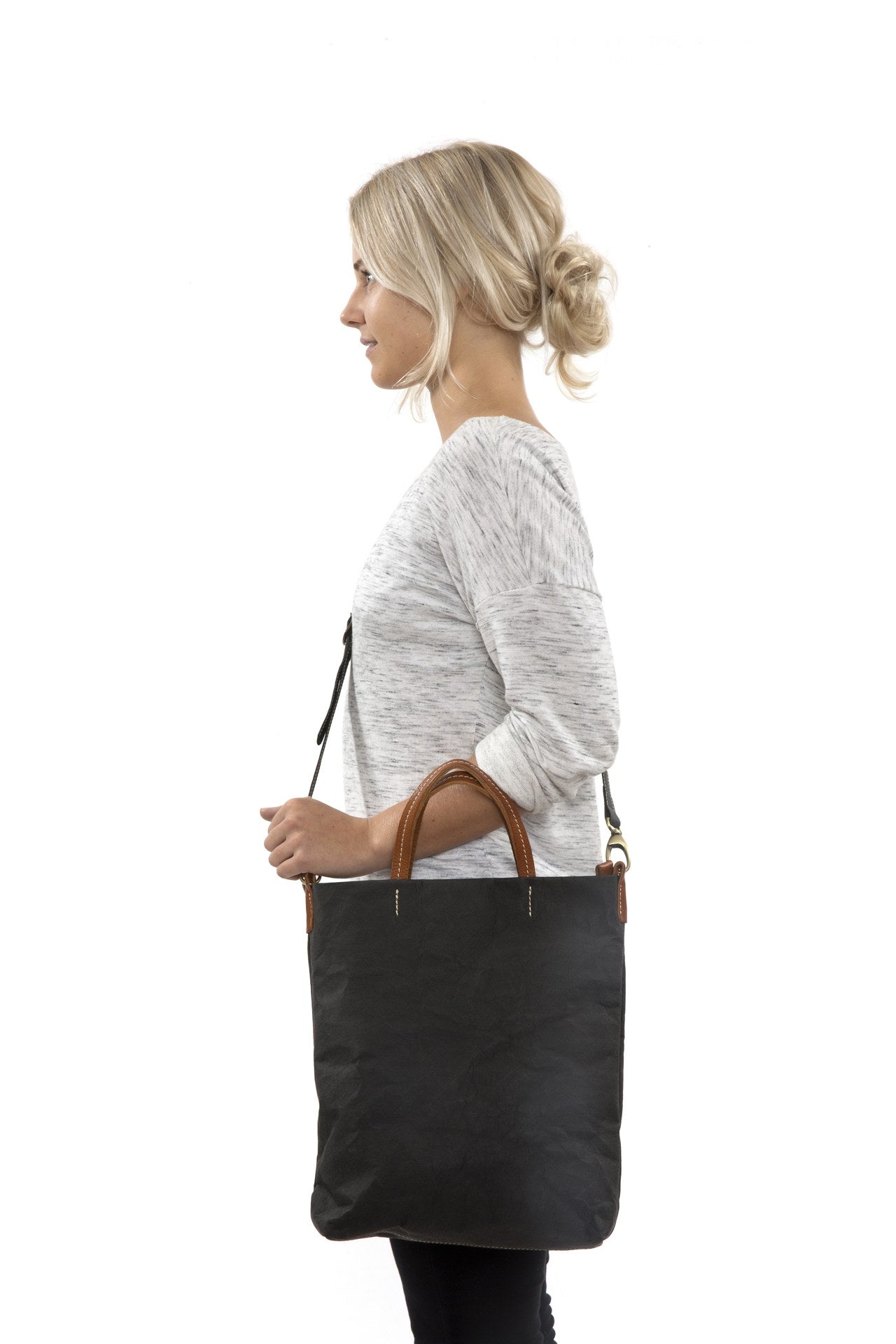 A blonde woman is shown from the side wearing casual clothing. She wears a crossbody washable paper tote bag with top handles. It is black in colour.